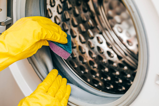 Ένας άντρας με κίτρινα γάντια καθαρίζει ένα βρώμικο, μουχλιασμένο λάστιχο σε ένα πλυντήριο. Μουλάρι, χώμα, λέπια στο πλυντήριο. Περιοδική συντήρηση οικιακών συσκευών. - Φωτογραφία, εικόνα