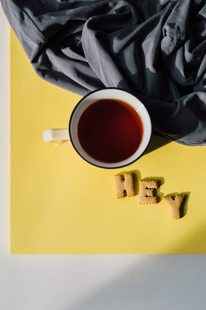 フラットレイアウト組成:グレーの繊維と明るい黄色のテーブルの上にお茶とビスケットのカップ。朝の動機、朝食のルーチン、またはリラックスした休憩の概念。2021色の傾向:黄色と灰色。トップ表示 - 写真・画像