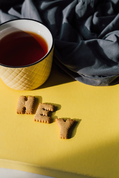 Egy csésze tea és keksz az élénk sárga asztalon szürke textilrel. Reggeli motiváció, reggeli rutin, lassú életmód vagy pihentető szünet koncepció. 2021 szín trendek: sárga és szürke. Felülnézet - Fotó, kép