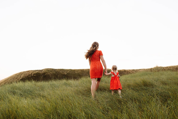 若い美しい母と娘が夏休みに自然の中を歩いています。夕日の時間に畑で遊んでいるママと女の子。母の日。友好的な家族の概念. - 写真・画像