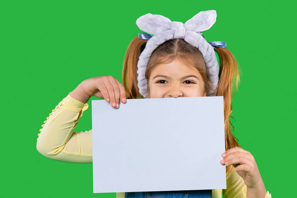 A preschool girl with a bandage on her head holding a white sheet for your advertisement on a green background. - Foto, afbeelding