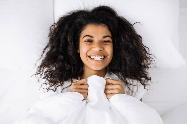 W weekendy rano. Widok góry portret atrakcyjnej dziewczyny relaksującej się w łóżku i uśmiechniętej. Zadowolona Afroamerykanka budzi się rano, czując się świeżo i radośnie, w weekendy w domu - Zdjęcie, obraz