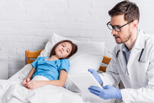 médico apuntando a la tableta digital mientras habla con un niño enfermo acostado en la cama - Foto, imagen