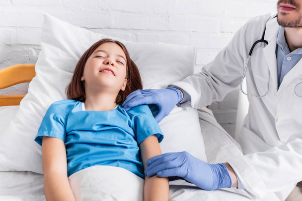 pediatrician examining diseased girl lying in bed with closed eyes - 写真・画像