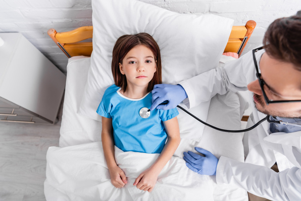 shora pohled na dívku ležící v posteli, zatímco lékař ji zkoumá stetoskopem - Fotografie, Obrázek