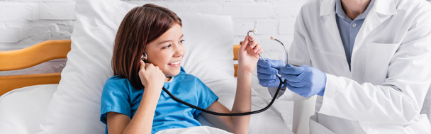 doctor showing stethoscope to cheerful girl in hospital bed, banner - Fotó, kép