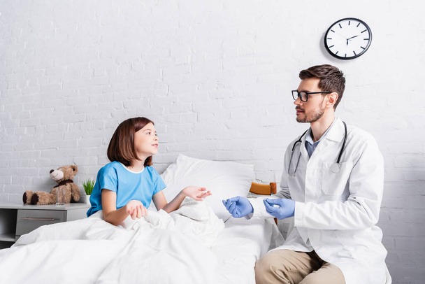 pediatrician in eyeglasses and girl in bed gesturing while talking in hospital - Foto, Imagen