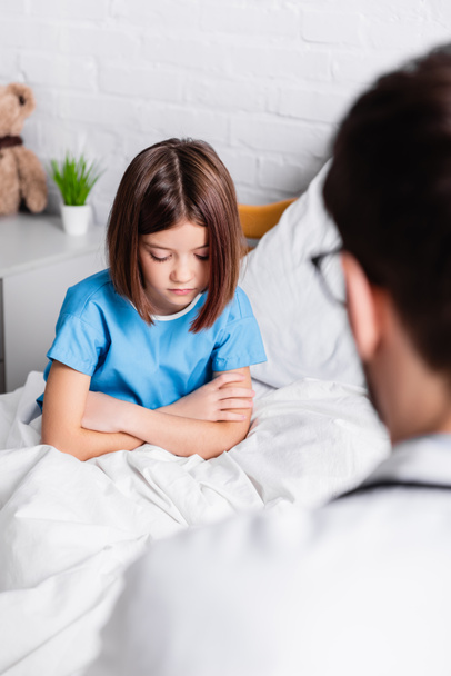 αναστατωμένο κορίτσι με σταυρωμένα χέρια στο κρεβάτι του νοσοκομείου κοντά στο γιατρό σε θολή πρώτο πλάνο - Φωτογραφία, εικόνα