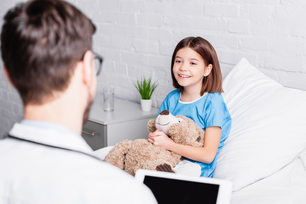 happy girl with teddy bear looking at pediatrician with digital tablet on blurred foreground - Photo, Image