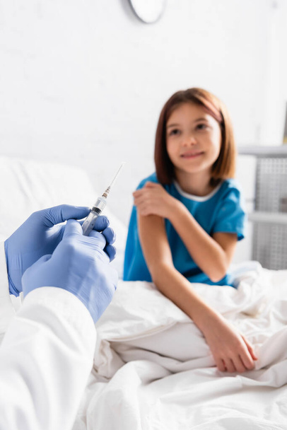 pediatrician holding syringe near smiling child sitting on bed - Photo, Image