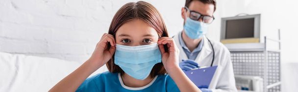 больной ребенок чинит медицинскую маску, глядя в камеру рядом с доктором, пишущим диагноз на размытом фоне, баннер - Фото, изображение