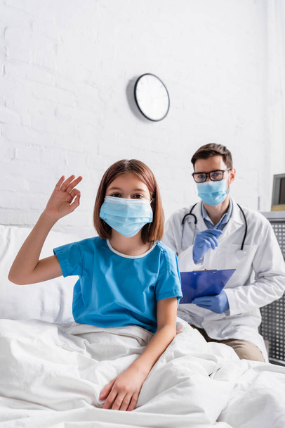 child in medical mask showing okay gesture near doctor with clipboard on blurred background - Fotó, kép