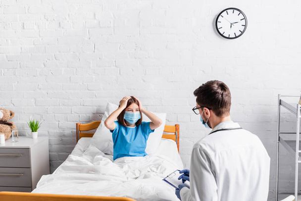 больная девушка в медицинской маске касается головы, когда страдает от головной боли рядом с доктором, пишущим на планшете - Фото, изображение