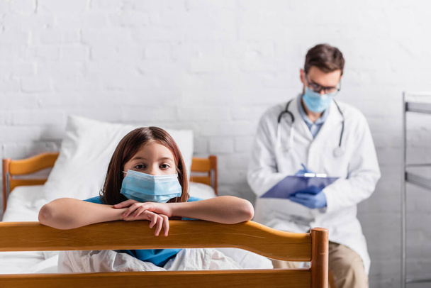 chica enferma en máscara médica mirando a la cámara cerca del médico escribiendo en el portapapeles sobre fondo borroso - Foto, imagen