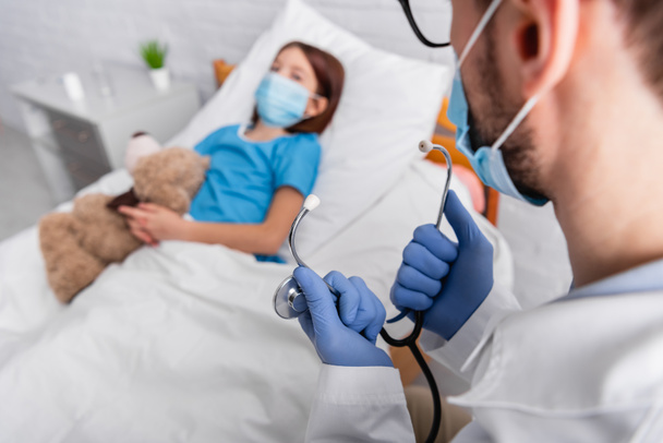 doctor sosteniendo estetoscopio cerca de niño enfermo en máscara médica acostado en la cama con oso de peluche sobre fondo borroso - Foto, Imagen