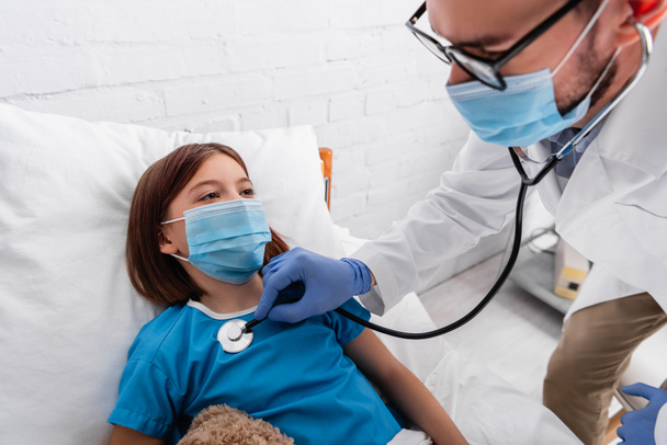 pediatrician in medical mask examining sick girl with stethoscope in hospital - Fotoğraf, Görsel