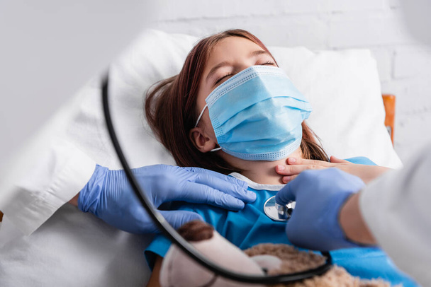 doctor examining girl, lying in medical mask with closed eyes, with stethoscope, blurred foreground - Foto, afbeelding