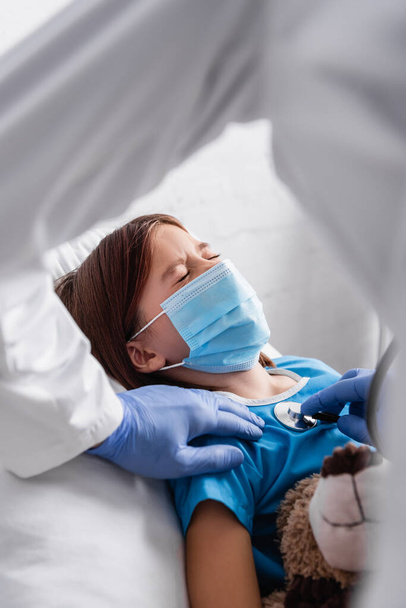 diseased girl in medical mask lying with closed eyes near doctor examining her with stethoscope, blurred foreground - Photo, image