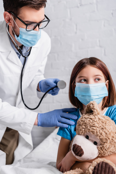 pediatrician with stethoscope near girl in medical mask sitting in bed with teddy bear - Photo, Image