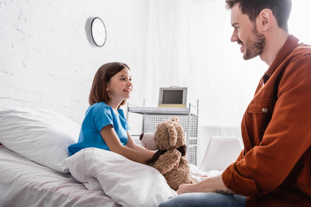 smiling girl sitting on hospital bed with teddy bear near happy dad, blurred foreground - Foto, imagen