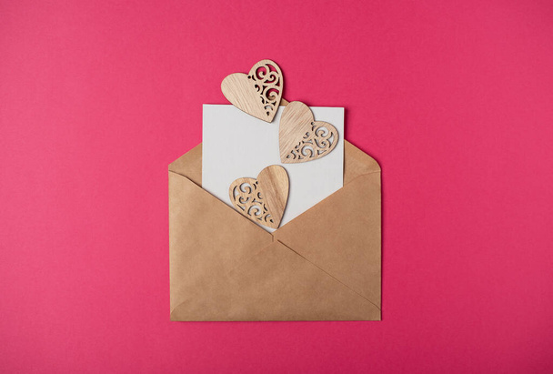 ホットピンクの背景に紙の空白のシートと木製の心を持つクラフト封筒。バレンタインデーのコンセプトのためのロマンチックな愛の手紙。テキストのスペース. - 写真・画像