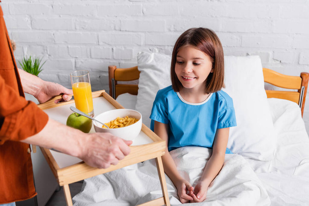 Mann hält Tablett mit Frühstück neben Tochter im Krankenhausbett, verschwommener Vordergrund - Foto, Bild