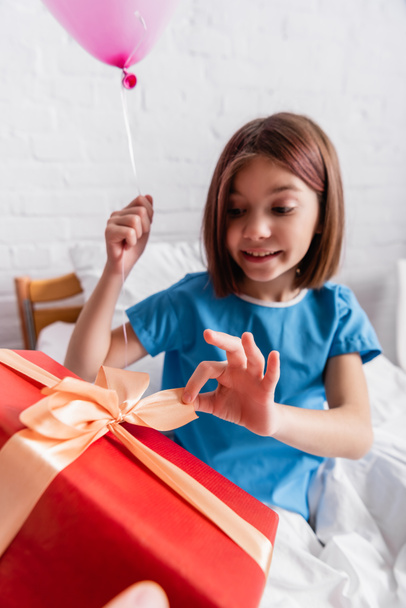 smiling girl opening birthday gift while holding festive balloon in hospital, blurred background - Фото, изображение