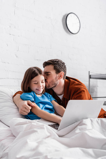happy father kissing cheerful daughter using laptop in hospital bed - Valokuva, kuva