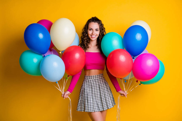 Portrait photo of pretty smiling girl wearing pink off-top checkered skirt holding air balloons in both hands isolated on vivid yellow color background - Fotó, kép