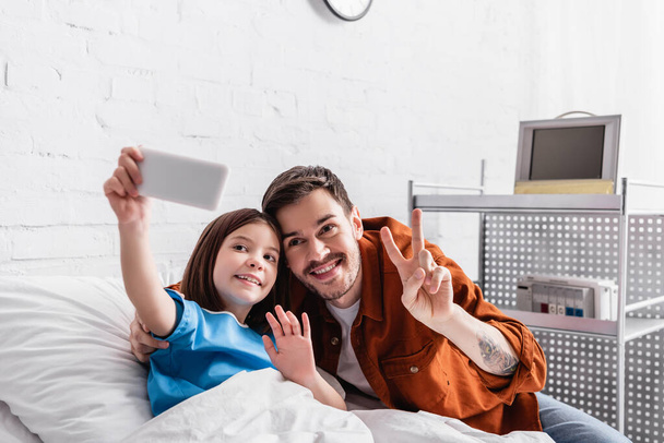 счастливый человек показывает победный жест рядом с дочерью размахивая рукой, делая селфи на смартфоне, размытый передний план - Фото, изображение