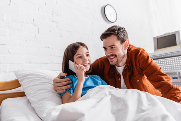 felice uomo abbraccio sorridente figlia parlando su smartphone in letto d'ospedale - Foto, immagini