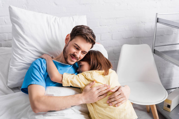 smiling man embracing daughter while lying in bed in hospital - Foto, Imagem