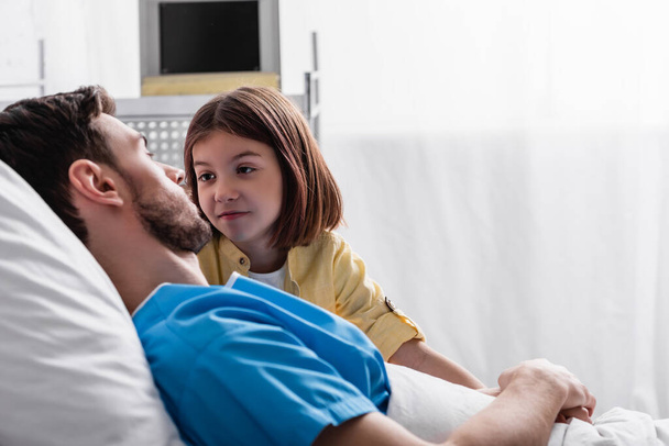 fille souriante regardant père couché dans le lit à l'hôpital, avant-plan flou - Photo, image