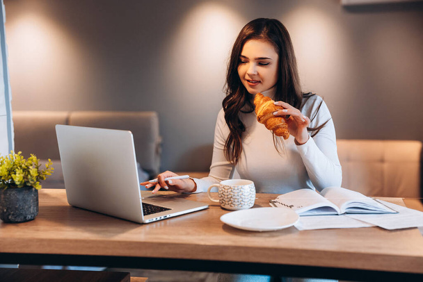 Freelance student woman using laptop while sitting at home. Young woman sitting in kitchen and working online on laptop. Pretty female drinking tea coffee while working - Foto, Imagem
