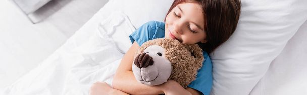 vue du dessus d'une fille souriante embrassant un ours en peluche pendant qu'elle dort à la clinique, bannière - Photo, image
