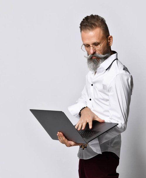 Studio portret hipstera w średnim wieku trzymającego laptopa i patrzącego w kamerę. - Zdjęcie, obraz