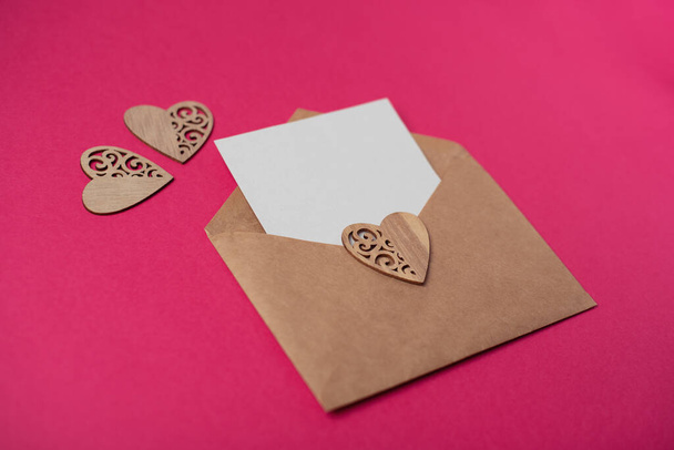 Craft envelope with a blank sheet of paper inside and wooden hearts on the hot pink background. Romantic love letter for the Valentine's day concept. Space for text. - Valokuva, kuva