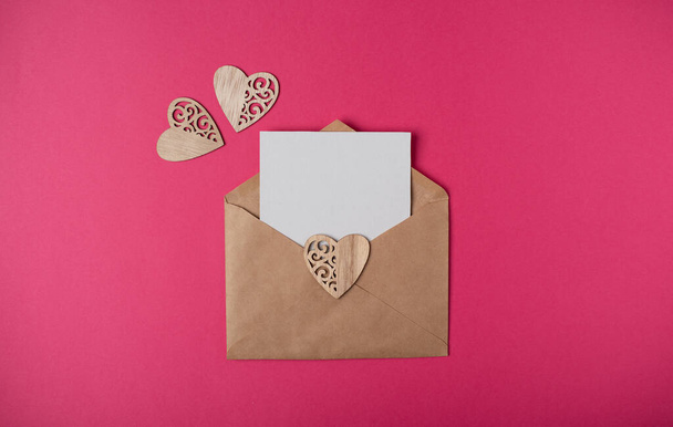 Craft envelope with a blank sheet of paper inside and wooden hearts on the hot pink background. Romantic love letter for the Valentine's day concept. Space for text. - Fotografie, Obrázek