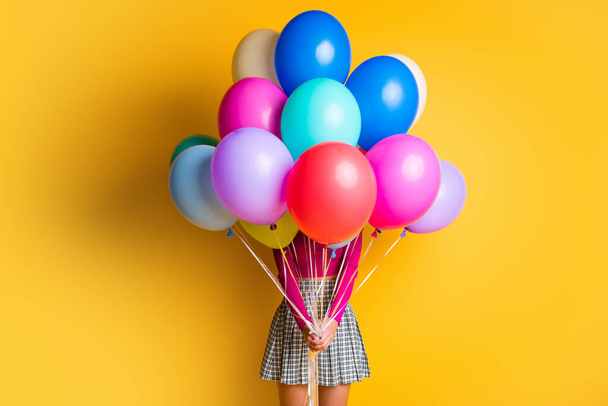 Portrét fotografie legrační dívka dělat překvapení skrývání tvář vedení barevné balónky na sobě ležérní oblečení izolované na žlutém pozadí - Fotografie, Obrázek