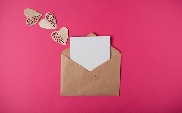 Craft envelope with a blank sheet of paper inside and wooden hearts on the hot pink background. Romantic love letter for the Valentine's day concept. Space for text. - Photo, Image