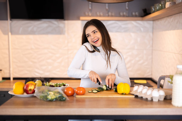 Красива дівчина ріже овочі для салату, розмовляє на мобільному телефоні і посміхається під час приготування їжі на кухні вдома
 - Фото, зображення