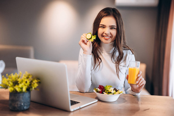 Joyful young woman drinking orange juice and eating salad from a bowl while standing on a kitchen and watching movie on laptop. - Foto, immagini