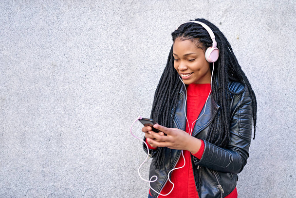 Un ritratto di una donna afro-americana sorridente con un telefono cellulare sta chiacchierando o ascoltando musica per le strade della città. I capelli sono intrecciati in dreadlocks. - Foto, immagini