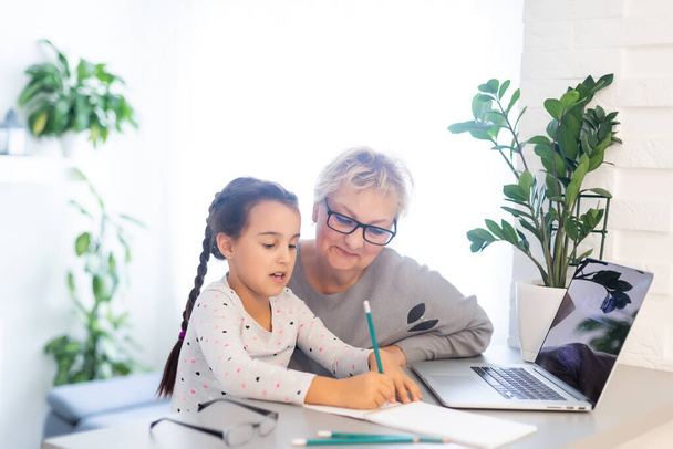 Mature grandmother helping child with homework at home. Satisfied old grandma helping her granddaughter studying in living room. Little girl writing on notebook with senior teacher sitting next to her - Photo, Image