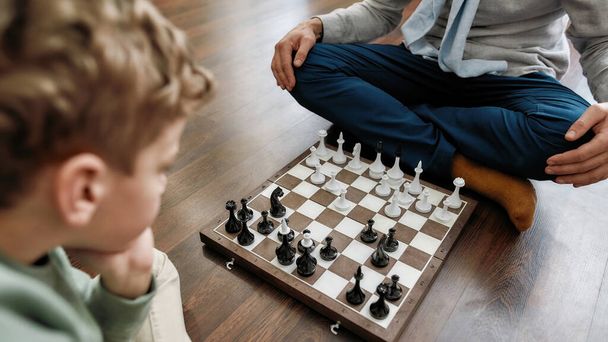 Intellectual game. Little thoughtful boy playing chess with grandfather while sitting on the floor in living room at home - 写真・画像