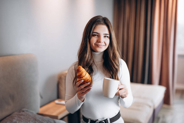 Appealing young girl is eating croissant and drinking coffee tea at home. Cute student is having coffee break - Fotografie, Obrázek