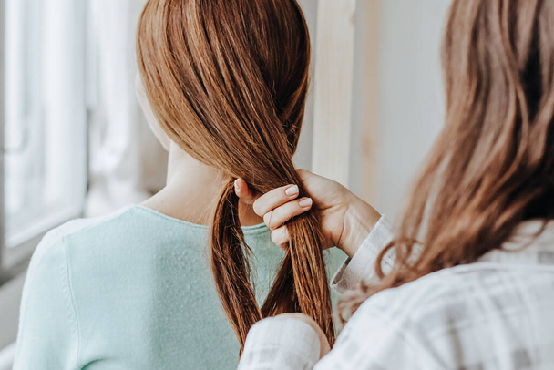 Two girls braid their hair at the window. Woman makes a braid to her friend. Hair weaving hairstyles. Girlfriend braids her hands with ringlets. Hair care - 写真・画像