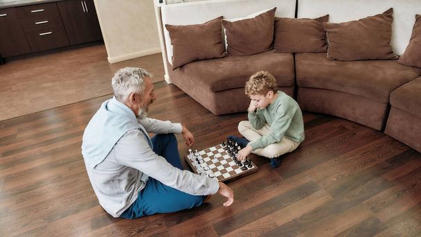 Abuelo enseñando a su nieto la mejor estrategia y tácticas de ajedrez mientras están sentados juntos en el suelo en la sala de estar, pasando tiempo en casa - Foto, Imagen