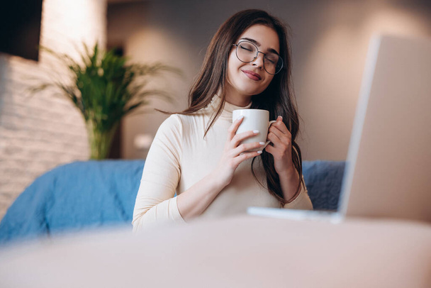 Krásná dívka v brýlích se těší šálek čaje kávy při práci na notebooku uvnitř. Žena na volné noze pracující z domova - Fotografie, Obrázek