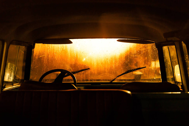 Interior del vehículo con volante vintage, asientos sin apoyacabezas sobre el fondo del parabrisas mojado, limpiaparabrisas, gotas de lluvia y luz de calle por la noche. Clima lluvioso. - Foto, Imagen